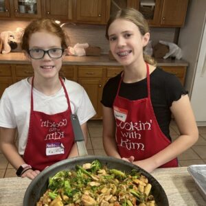 Two girls in the kitchen with a large pan of cooked food.