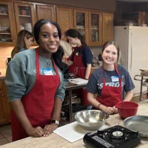 Two women cooking together in a cozy kitchen, wearing red aprons.