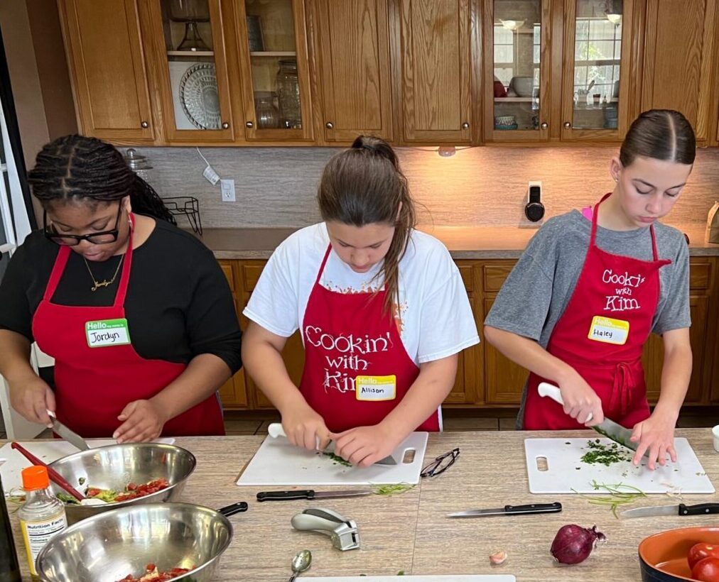 Three children wearing red aprons preparing food together at a kitchen counter.