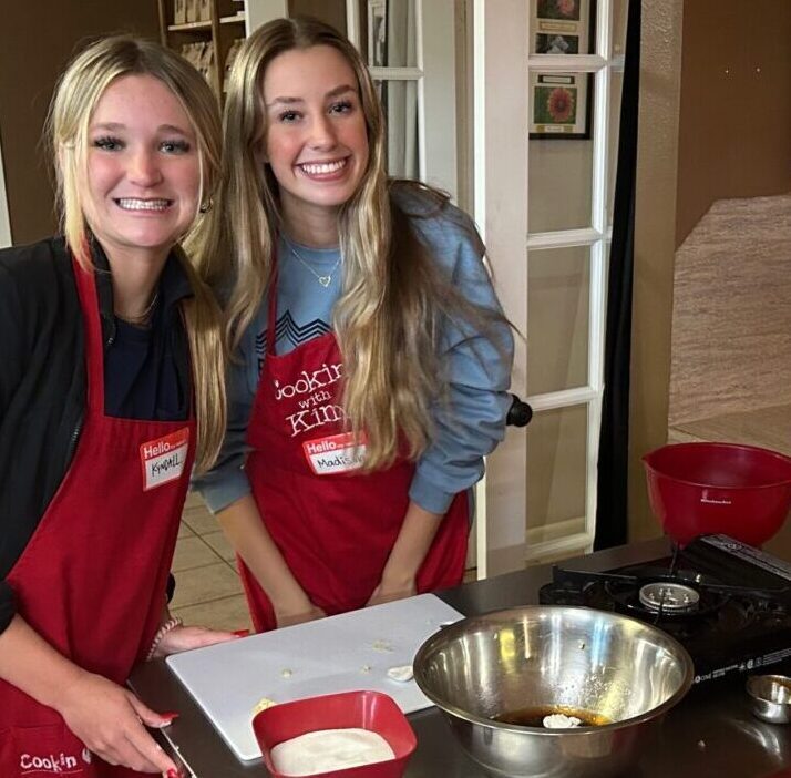 Three young women in red aprons smiling behind a kitchen counter with cooking supplies.