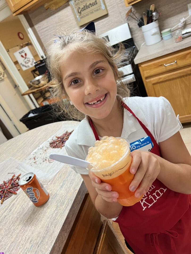 A smiling girl holding a colorful drink with whipped cream in a kitchen.